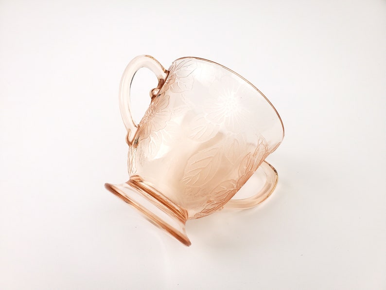May include: A pink glass creamer with a floral design and a handle. The creamer has a pedestal base.
