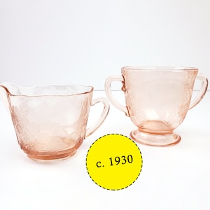 May include: A vintage pink glass creamer and sugar bowl set with a floral design. The set is dated c. 1930.
