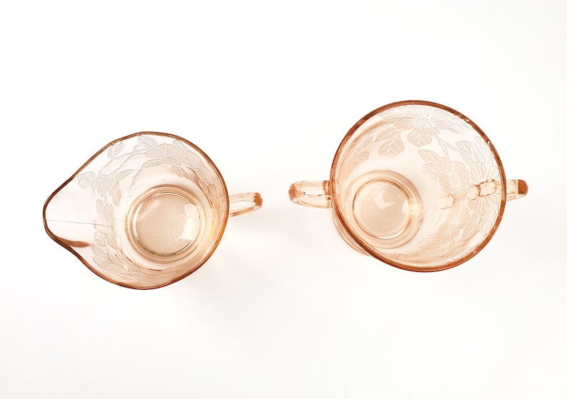 May include: A set of two vintage glass creamer and sugar bowls with a floral design. The bowls are made of pink glass and have a delicate etched pattern.