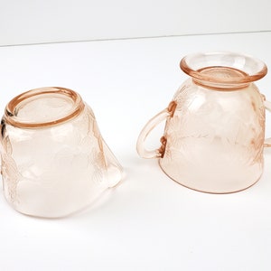 May include: A vintage pink glass creamer and sugar bowl set with a floral design. The creamer has a handle and a spout, while the sugar bowl has two handles.