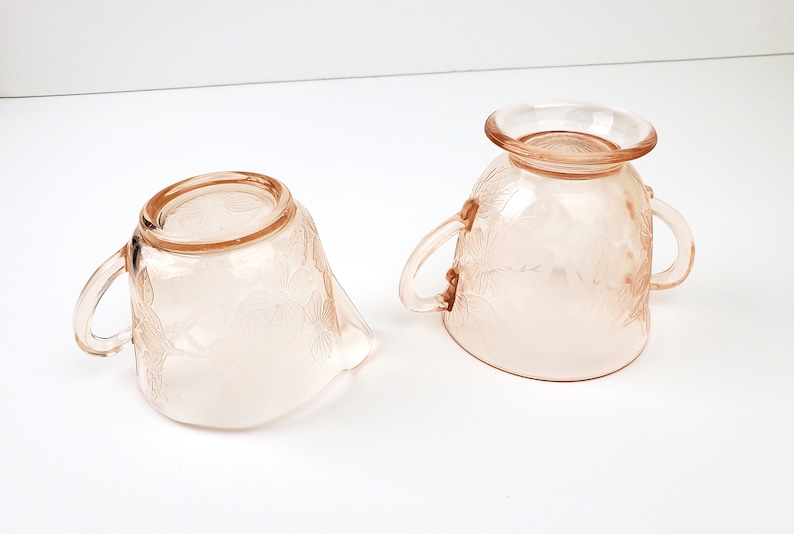 May include: A vintage pink glass creamer and sugar bowl set with a floral design. The creamer has a handle and a spout, while the sugar bowl has two handles.