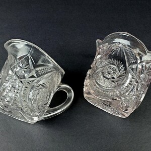May include: Two clear glass jugs with intricate cut glass designs. The jugs have a rounded body and a handle.