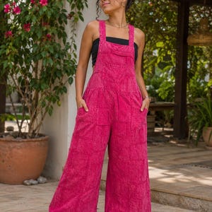 May include: A pair of bright pink denim overalls featuring a patchwork design with wide legs, a front bib pocket, and side pockets. The garment has a distressed, acid-wash texture and is worn over a simple black tube top with white sneakers.