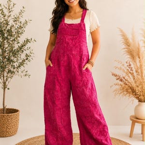 May include: A pair of bright magenta denim overalls featuring a relaxed, wide-leg fit with a patch-style construction, a front bib pocket, and two side pockets, worn over a short-sleeved, cream-colored ribbed top with a lettuce-edge neckline.