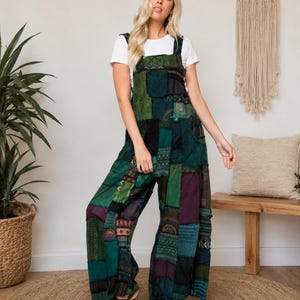 May include: A pair of patchwork overalls featuring a variety of green, teal, and purple fabric panels with diverse patterns. The overalls have a square neckline, wide legs, and are worn over a plain white short-sleeved t-shirt with tan sandals.