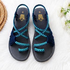 May include: A pair of dark gray sandals featuring a toe loop and crisscrossing braided straps in navy blue and turquoise. The footbed is dark gray with the number 6 and a gold logo printed at the heel. The sandals have a slingback strap design.