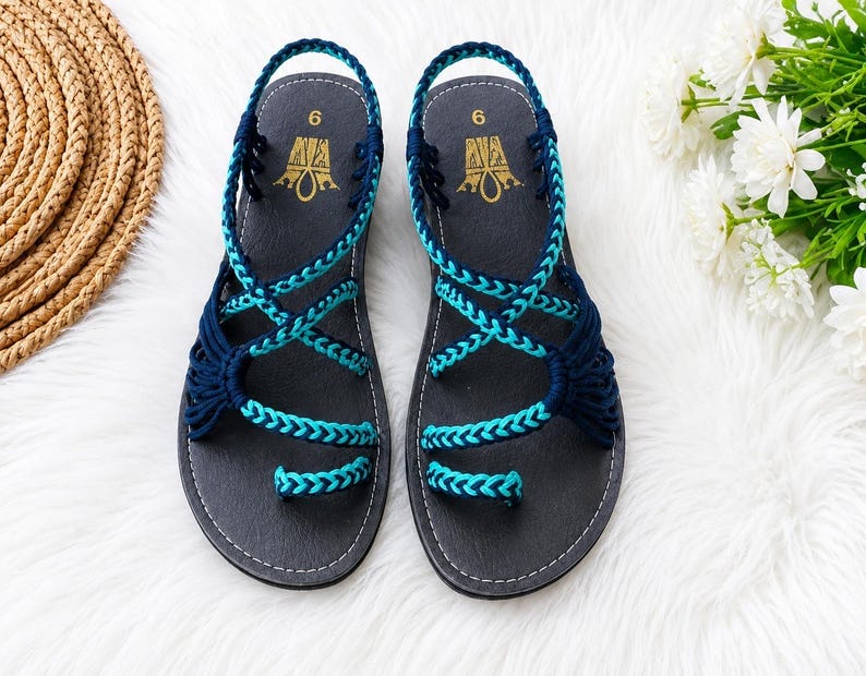 May include: A pair of dark gray sandals featuring a toe loop and crisscrossing braided straps in navy blue and turquoise. The footbed is dark gray with the number 6 and a gold logo printed at the heel. The sandals have a slingback strap design.