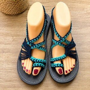 May include: A pair of flat sandals featuring black soles with white stitching and intricate braided straps in alternating dark navy blue and bright turquoise cord. The design includes a toe loop and multiple crisscrossing straps that secure the foot.