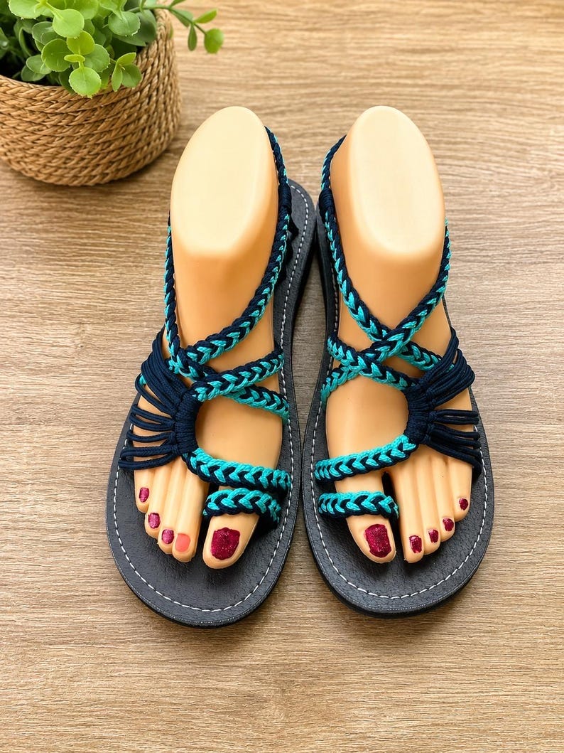 May include: A pair of flat sandals featuring black soles with white stitching and intricate braided straps in alternating dark navy blue and bright turquoise cord. The design includes a toe loop and multiple crisscrossing straps that secure the foot.