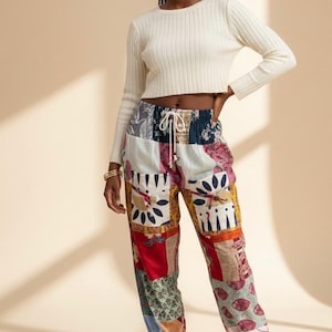 May include: A woman wearing a cream-colored, ribbed long-sleeve crop top and patchwork pants. The pants feature a variety of colorful fabric patches with floral and geometric patterns. The pants have a drawstring waist and elastic cuffs.