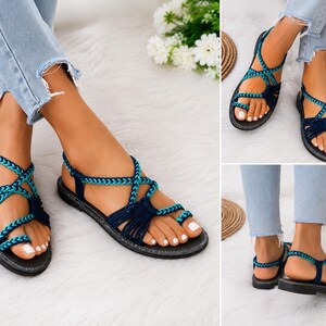 May include: Flat sandals featuring a dark blue and teal braided strap design with a toe loop and an adjustable ankle strap. The footwear has a black sole and a multi-strap construction that secures the foot across the top and around the heel.