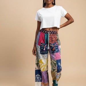 May include: A woman wearing a white crop top and colorful patchwork pants. The pants feature a variety of patterns and colors, including blue, pink, green, and yellow. The pants have a drawstring waist. The woman is also wearing white sandals.