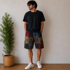 May include: A person wearing a black t-shirt and patchwork shorts in shades of red, blue, green, and brown. The person is wearing white sneakers and sunglasses. A plant in a woven basket is to the left.