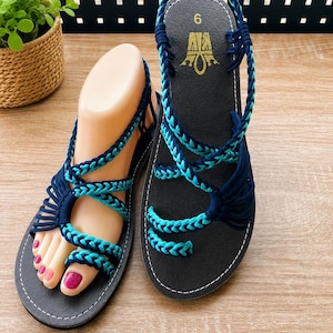 May include: A pair of flat sandals featuring black soles and intricate braided straps in dark blue and turquoise. The straps wrap around the foot and include a toe loop design. The footbed displays the number 6 and a gold logo. The sandals are constructed with multiple cord-like strands that secure the foot to the base.