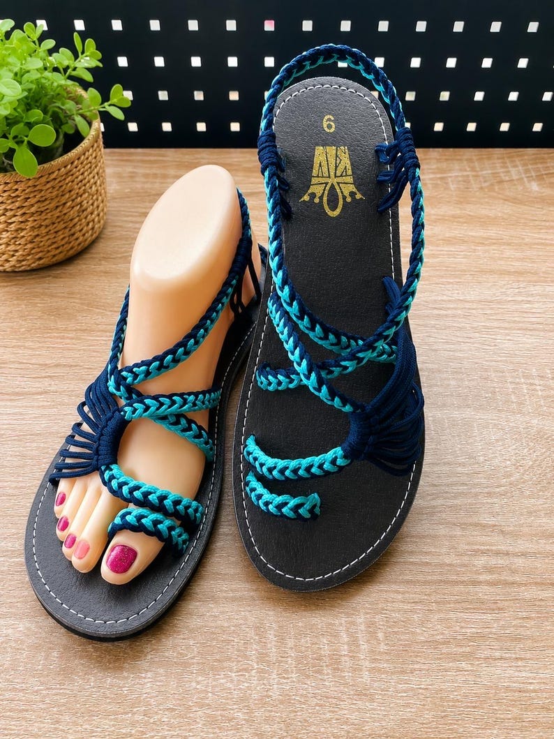 May include: A pair of flat sandals featuring black soles and intricate braided straps in dark blue and turquoise. The straps wrap around the foot and include a toe loop design. The footbed displays the number 6 and a gold logo. The sandals are constructed with multiple cord-like strands that secure the foot to the base.