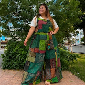 May include: A sleeveless jumpsuit featuring a patchwork design with various shades of green, brown, and red fabric squares. The garment has wide legs and is worn over a plain white short-sleeved t-shirt. The fabric displays diverse textures and patterns.