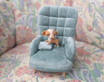 Könnte beinhalten: Ein kleiner, hellblauer Sessel mit einem gesteppten Rücken und einem weichen Kissen. Eine kleine, braun-weiße Hundefigur sitzt auf dem Kissen.