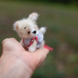 May include: A small, white and brown crocheted dog wearing a red and blue dress. The dog is sitting in the palm of a person's hand.