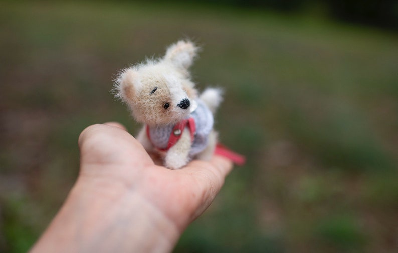 May include: A small, white and brown crocheted dog wearing a red and blue dress. The dog is sitting in the palm of a person's hand.