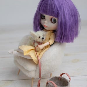 May include: A doll with purple hair and a yellow dress sits in a fluffy white chair, holding a small white dog on a pink leash. A small woven basket with pink leather handles sits nearby. The scene evokes a whimsical, miniature world.