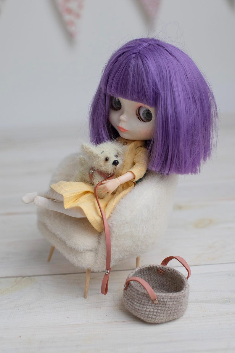 May include: A doll with purple hair and a yellow dress sits in a fluffy white chair, holding a small white dog on a pink leash. A small woven basket with pink leather handles sits nearby. The scene evokes a whimsical, miniature world.