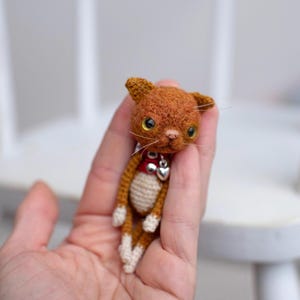 May include: A small, handcrafted ginger cat figurine made of textured yarn. The cat has a cream-coloured belly, white paws, and large, glossy yellow eyes. It wears a red collar featuring a small, shiny silver heart-shaped charm around its neck.