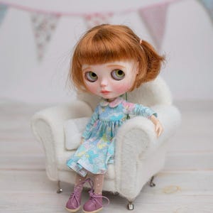 Könnte beinhalten: Puppe mit rotem Haar und grünen Augen, trägt ein blaues Blumenkleid und rosa Schuhe, sitzt in einem weißen, flauschigen Sessel. Das Kleid hat einen hellblauen Hintergrund mit bunten Blumenmustern. Im Hintergrund befindet sich ein dekoratives Banner.