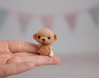 May include: A tiny, light brown teddy bear figurine with small, dark eyes and ears. The bear is held in a person's hand, highlighting its small size. The bear has a pearl necklace and a white bow. The background is blurred with pastel colours.