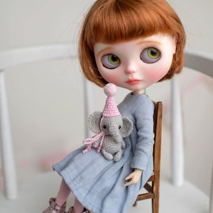 Puede incluir: Muñeca con cabello castaño rojizo y ojos verdes, vestida con un vestido azul claro y zapatos rosas. La muñeca sostiene un pequeño elefante gris tejido con un gorro de fiesta y una bufanda rosas. La muñeca está sentada en una silla de madera.