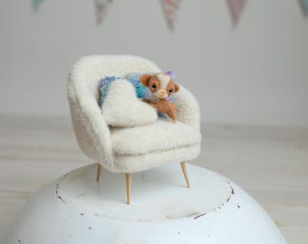 Könnte beinhalten: Ein cremefarbener Miniatur-Sessel mit Holzbeinen. Eine kleine, braun-weiße Hunde-Figur liegt auf dem Stuhl, eingebettet in ein blau-weißes Kissen. Der Stuhl steht auf einer weißen, abgerundeten Oberfläche.