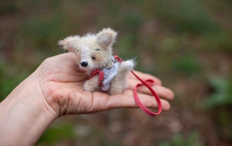 May include: A small, white and brown knitted dog toy with a red leash. The dog is wearing a pink and white knitted vest.