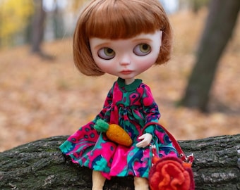 May include: A doll with auburn hair and large eyes wearing a colorful dress with a green collar, red shoes, and holding a carrot. A small red purse sits beside the doll. The doll is sitting on a tree trunk in a forest setting.