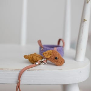 以下が含まれることがあります： 茶色の体とタン色の頭を持つ手作りのミニチュアかぎ針編みの犬。ピンク色の革のリードが付いています。革のハンドルが付いた小さな紫色のバスケットが背景にあります。アイテムは白い木の表面にあります。