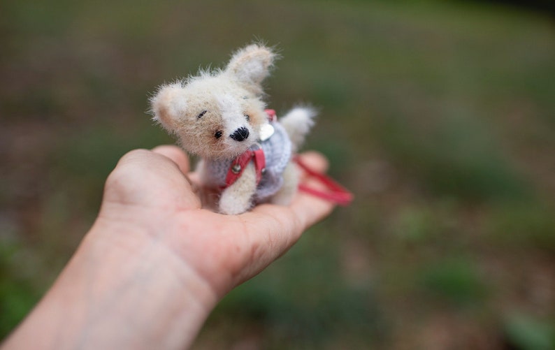 May include: A small, white and brown knitted toy dog wearing a red and gray sweater. The dog is being held in a person's hand.