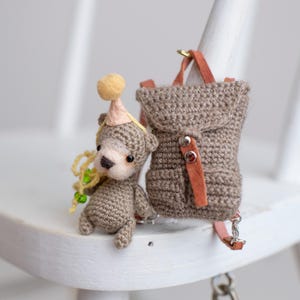 Puede incluir: Una figura de oso de ganchillo con un gorro de fiesta, junto a una mochila a juego. El oso es de color marrón claro con un hocico color crema y un pompón amarillo en su gorro. La mochila tiene correas de cuero y cierres metálicos.