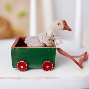 以下が含まれることがあります： 白いガチョウのぬいぐるみが、赤い車輪のついた緑色の木製ワゴンに乗っています。ガチョウは、かぎ針編みのセーターを着ています。ワゴンには、茶色の革のストラップが付いています。