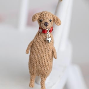 Puede incluir: Pequeña figura de perro tejido en tonos marrón. Tiene un collar rojo con un dije de corazón plateado. El perro tiene ojos pequeños y oscuros y una nariz marrón. La figura está sobre una superficie blanca.