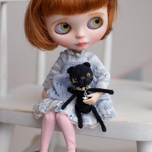 May include: A doll with red hair and green eyes, wearing a blue and white dress and pink leggings, holds a small black crocheted cat with yellow eyes and a silver star pendant. The doll is seated on a white chair.