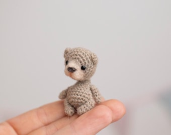 May include: A miniature crocheted teddy bear in shades of grey and beige. The tiny bear has a detailed, textured appearance, with small black eyes and a light brown nose. It is sitting on a person's hand.