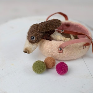 May include: A handcrafted miniature dachshund dog made of felt, nestled in a light pink felt basket with leather handles. Three small felt balls in green, orange, and pink are in front of the basket. The dog has brown and white fur.