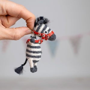 May include: A handmade crochet zebra figurine with black and white stripes, a red harness, and a black tail. The zebra is held up by a hand against a blurred background with colorful pennant flags.