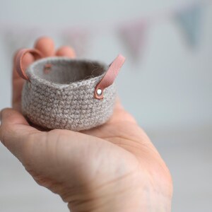 May include: A small, hand-crocheted basket in a neutral beige color. The basket has a rounded shape with a light pink leather handle attached with a silver rivet. The basket is held in a person's hand.