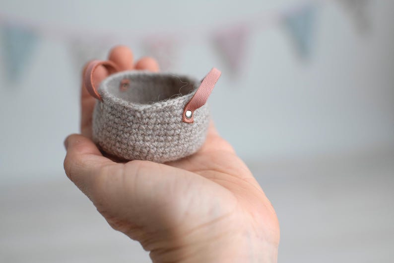 May include: A small, hand-crocheted basket in a neutral beige color. The basket has a rounded shape with a light pink leather handle attached with a silver rivet. The basket is held in a person's hand.