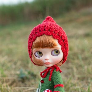 May include: A doll with large eyes and ginger hair wearing a red crocheted bonnet with a tie under the chin. The doll is dressed in a green knitted cardigan featuring red stripes on the sleeves and a small red and white mushroom accessory.