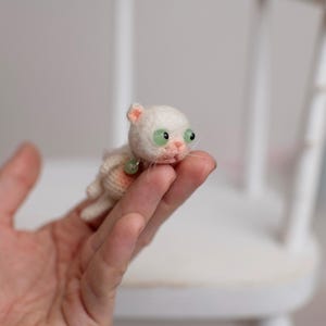 May include: A miniature white crocheted cat figurine with large green eyes, a small pink nose, and a tiny green bead collar. The small stuffed animal features a textured yarn surface and is crafted with delicate details including whiskers.