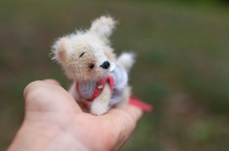 May include: A small, crocheted dog toy with white and brown fur, wearing a red and blue vest. The toy is being held in a person's hand.