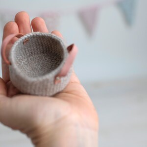 May include: A small, hand-knitted basket in a neutral gray color, featuring two pink leather handles. The basket is held in a hand, showcasing its size and texture. The background is blurred, with colorful bunting.