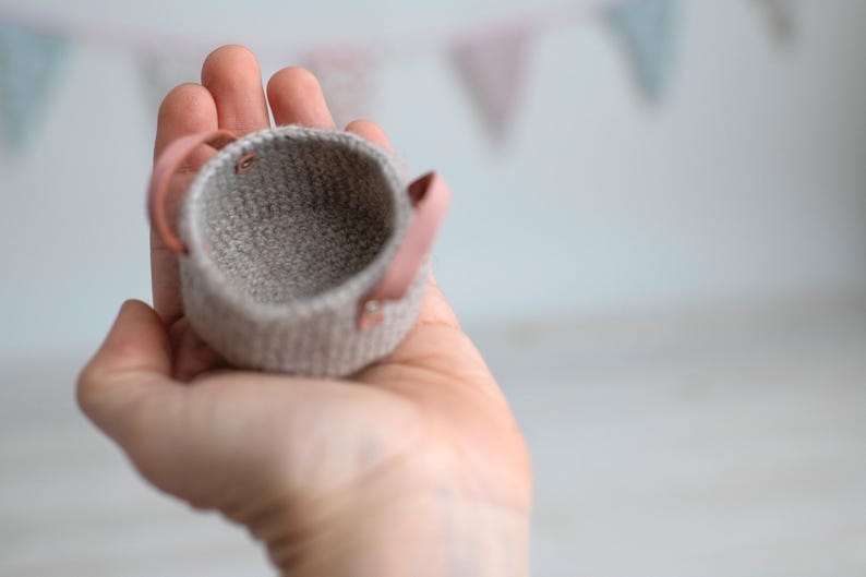 May include: A small, hand-knitted basket in a neutral gray color, featuring two pink leather handles. The basket is held in a hand, showcasing its size and texture. The background is blurred, with colorful bunting.