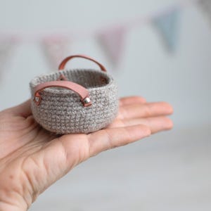 May include: A small, crocheted basket in a neutral beige color, held in a hand. The basket has two pink leather handles attached with silver rivets. The background is blurred with pastel-colored bunting.