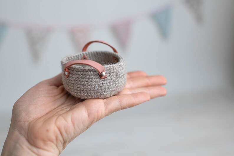 May include: A small, crocheted basket in a neutral beige color, held in a hand. The basket has two pink leather handles attached with silver rivets. The background is blurred with pastel-colored bunting.